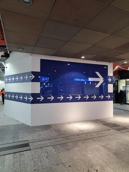 Weisse Zwischenwand in einer Unterfürung mit zwei blauen Streifen mit nach rechts zeigenden weissen Pfeilen. Darüber ein riesiges blaues Schild mit einem grossen, weissen, nach rechts zeigendem Pfeil.